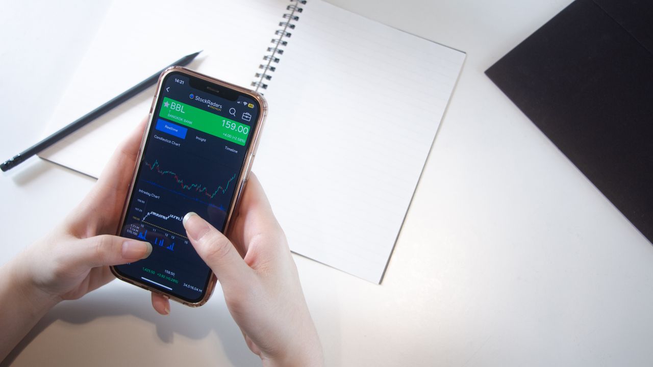 Gros plan sur des mains utilisant une application boursière sur smartphone, avec un carnet et un crayon sur un bureau.